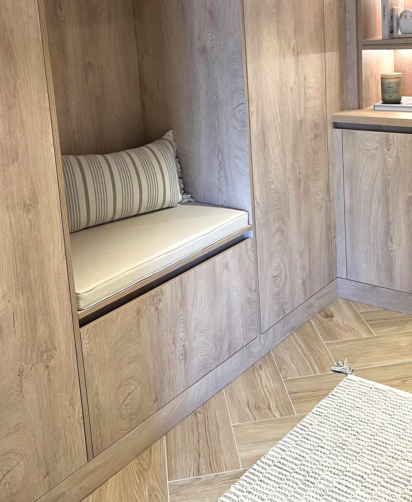Holiday home seating cushions made to measure. A light wood panelled living area with small kitchen counter and a seating nook between wooden cupboards. The ash wood storage bench has a light cream coloured herringbone weave fabric bench cushion, made to fit the shape and size of the seat. The thin foam seat pad has oatmeal coloured cotton fabric as the cover, with piping at the edges and a zip at the back so the bench cushion cover can be taken off for cleaned. Natural fibre, neutral colour