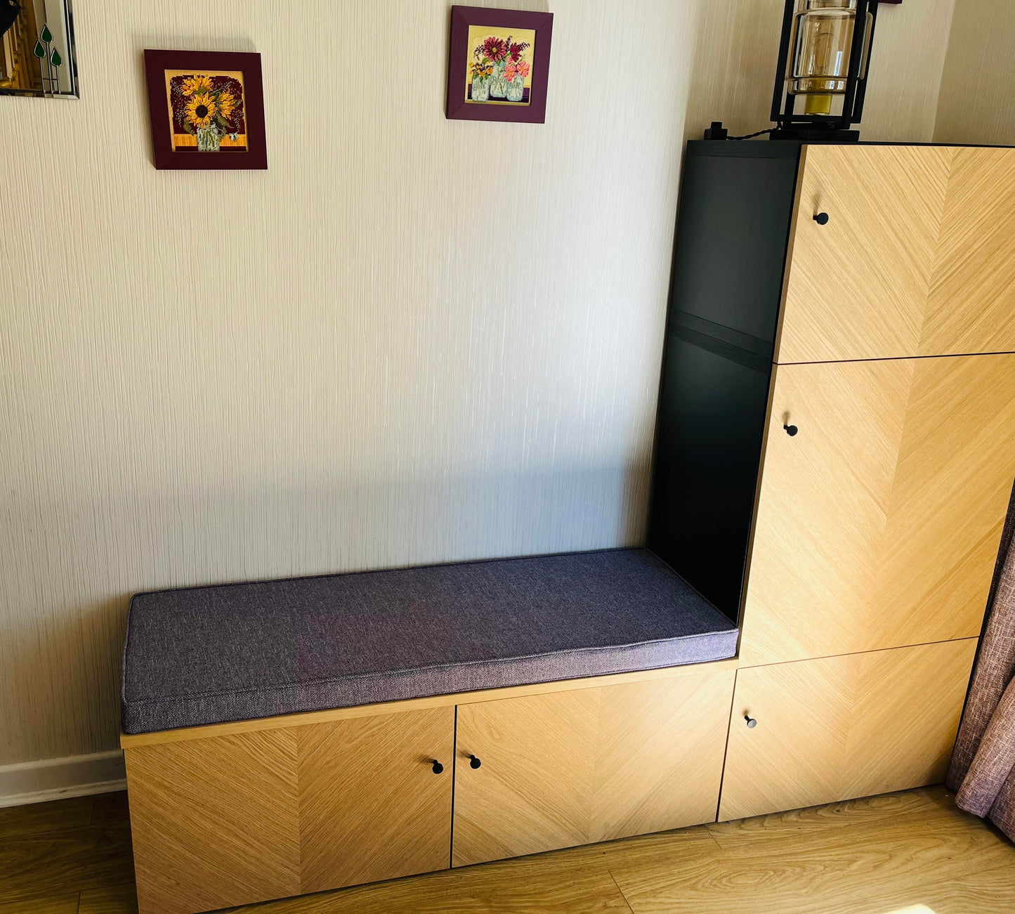 A bright living room with a modern, herringbone pattern wooden storage cupboard system with a storage bench area. The light wood cupboards have a thin firm foam bespoke bench cushion on top in a lavender purple, linen look woven upholstery fabric. The bench cushion cover has piping at the edges in a machine washable cord and a zip at the back long edge of the cushion so the cover can be removed for washing. Stain resistant, velcro straps, anti-slip. William Morris, Liberty fabric, Sophie Allport