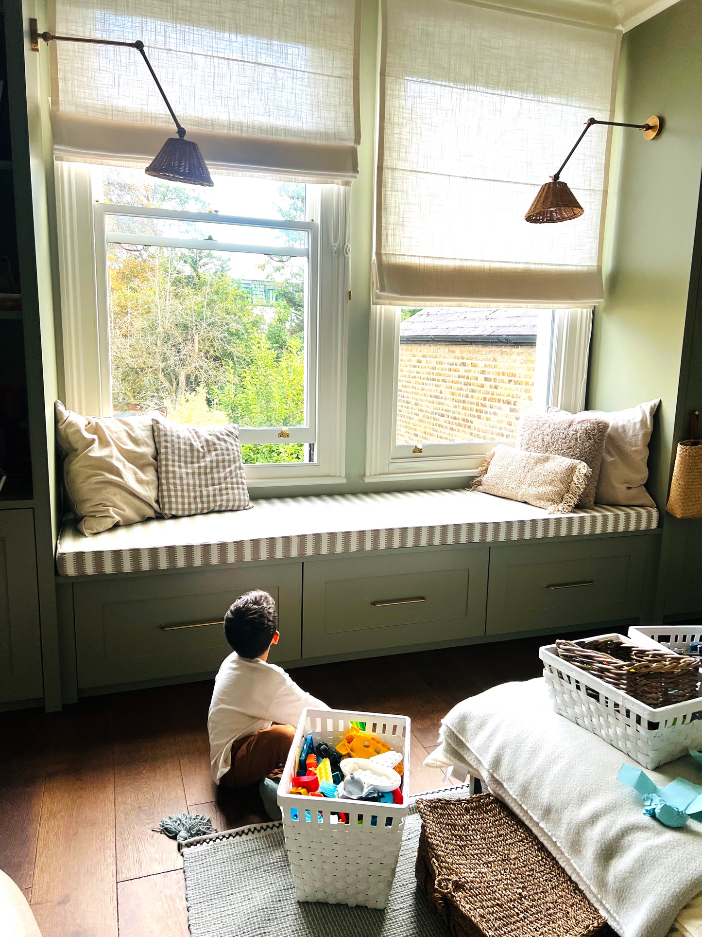 A brightly lit child's bedroom with sage coloured walls and wooden flooring. There is a wide wooden sash window with a window seating ledge. On top the the window seat is a made to measure foam bench cushion in a Harlequin stripe performance fabric. The beige and white geometric stripe bench cushion cover has piping at the edges and a zip at the back so it can be washed. The stripey bench cushion has been made to order in the UK by a small local company and is high quality. 