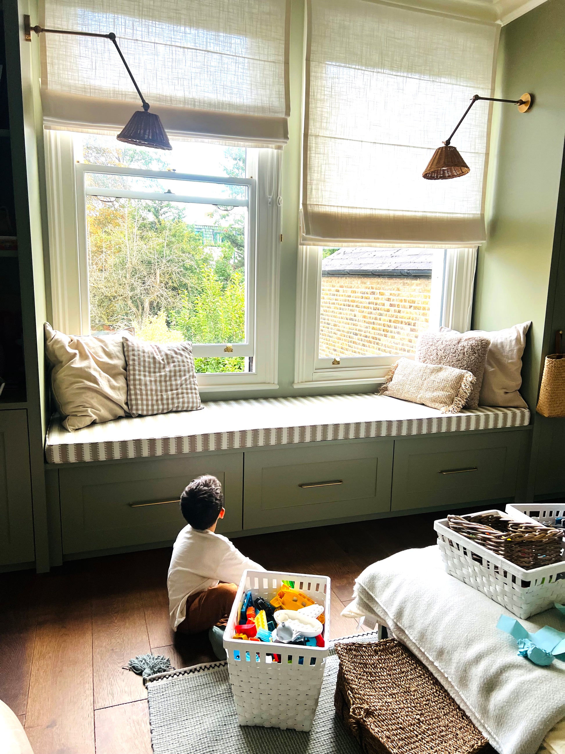 A brightly lit child's bedroom with sage coloured walls and wooden flooring. There is a wide wooden sash window with a window seating ledge. On top the the window seat is a made to measure foam bench cushion in a Harlequin stripe performance fabric. The beige and white geometric stripe bench cushion cover has piping at the edges and a zip at the back so it can be washed. The stripey bench cushion has been made to order in the UK by a small local company and is high quality. 
