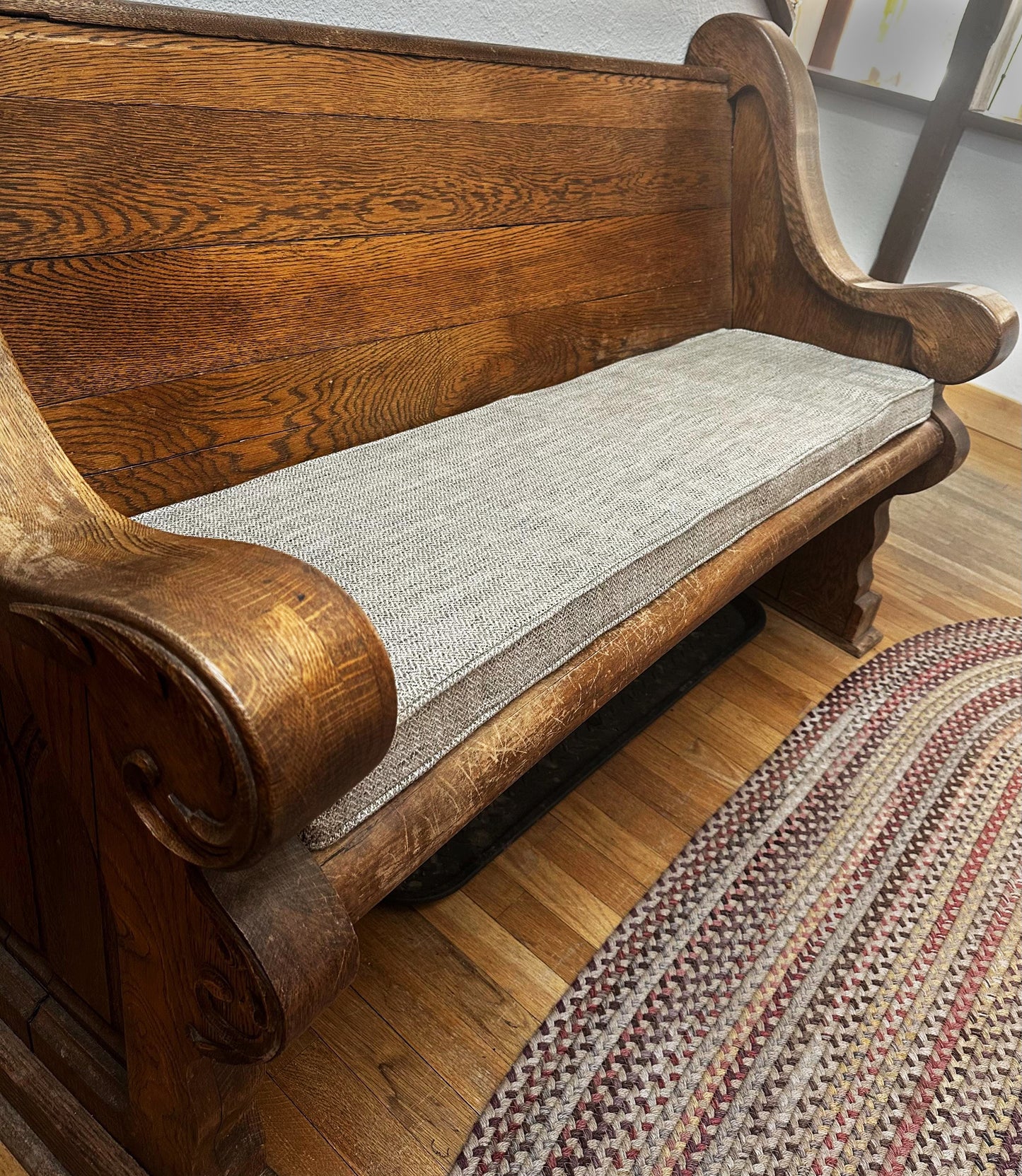 A thick, heavy looking oak wood bench with sturdy arms. A thin bench cushion on top of the church pew in a light brown herringbone patterned upholstery fabric with firm foam inside. Made to measure indoor bench cushion in patterned fabrics with stain resistant treatment. Pet friendly bench cushion, wipe clean window seat cushion with piped edges. Zippered bench cushion cover for dining banquette. Hanging back cushions. Overlocked finished seams, Ercol sofa cushions shaped edges, curved corners