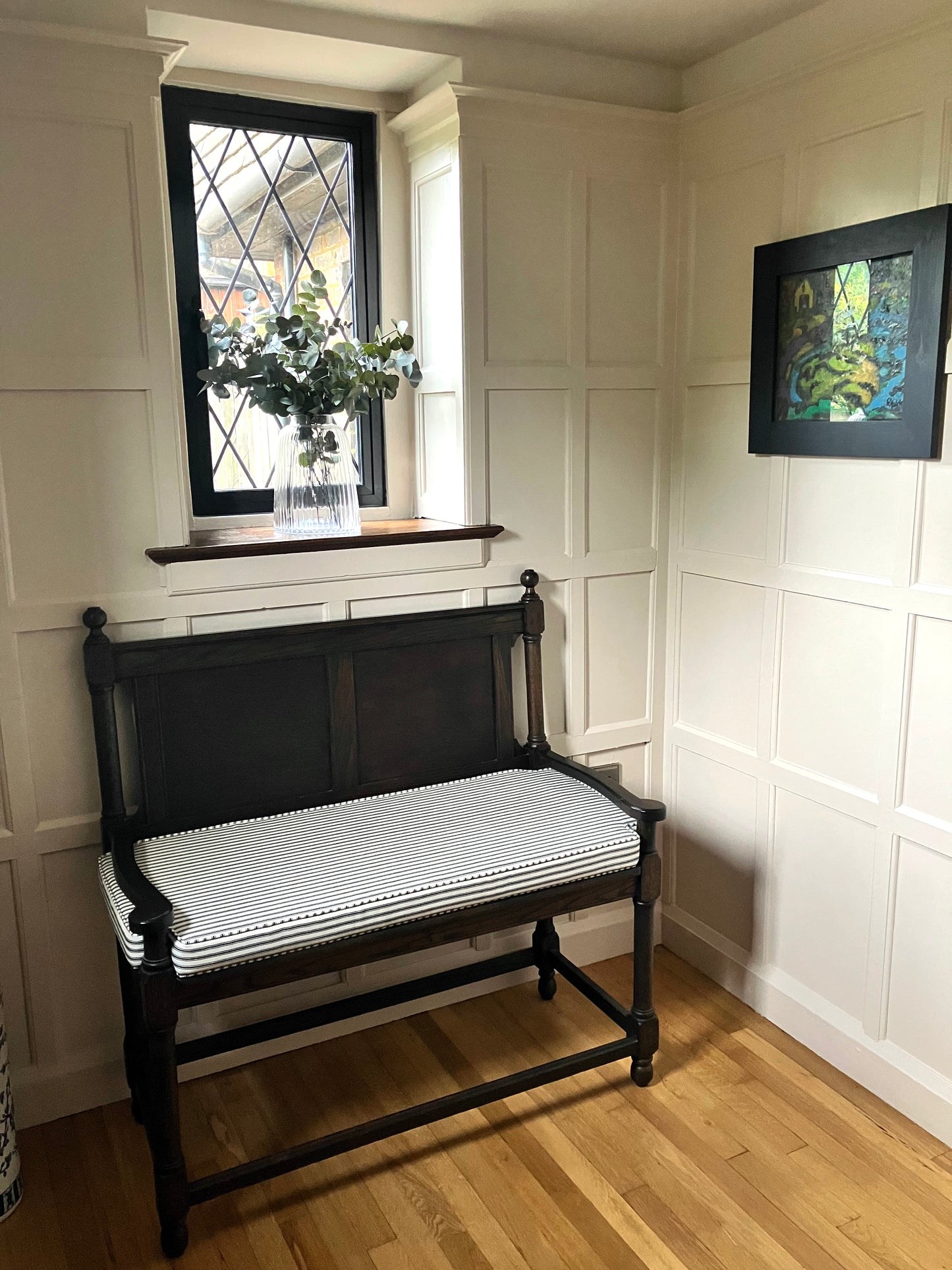 A classic wood panelled room corner painted in an off white Farrow and Ball colour. There is a dark wooden church pew bench that has a well made french ticking fabric bench cushion with cut outs for the bench arms. The cushion is a cotton fabric in a black and white ticking and has piped edges and a zip at the back of the cover. The custom shaped pew cushion has thin firm foam insert and piping at the edges. The stain resistant, striped fabric is a 100% cotton upholstery fabric made in the UK