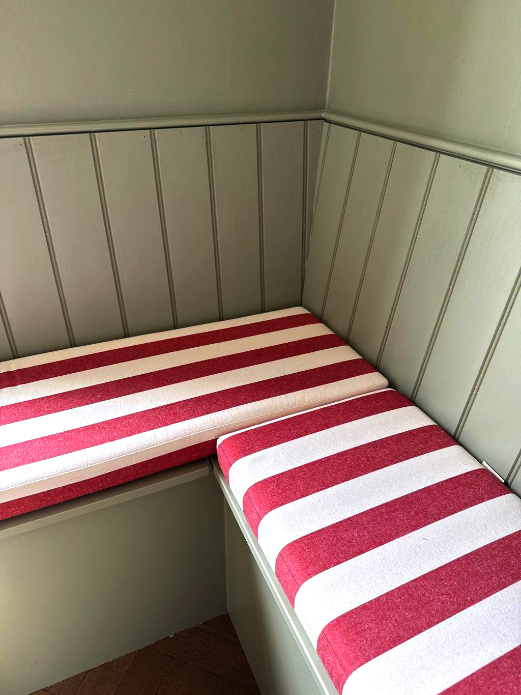 A corner bench seating unit in a kitchen dining room space. The wooden in built banquette has a wood panelled back rest and storage bench painted in a farrow and ball sage green colour. The firm foam bench cushions have thick deck chair style stripes in white and red. The cotton bench cushion cover is made to measure in an Ian Mankin ticking style fabric. The custom made bench cushion covers have zips at the back so they can be removed and have no piping on the edges for a modern kitchen style.