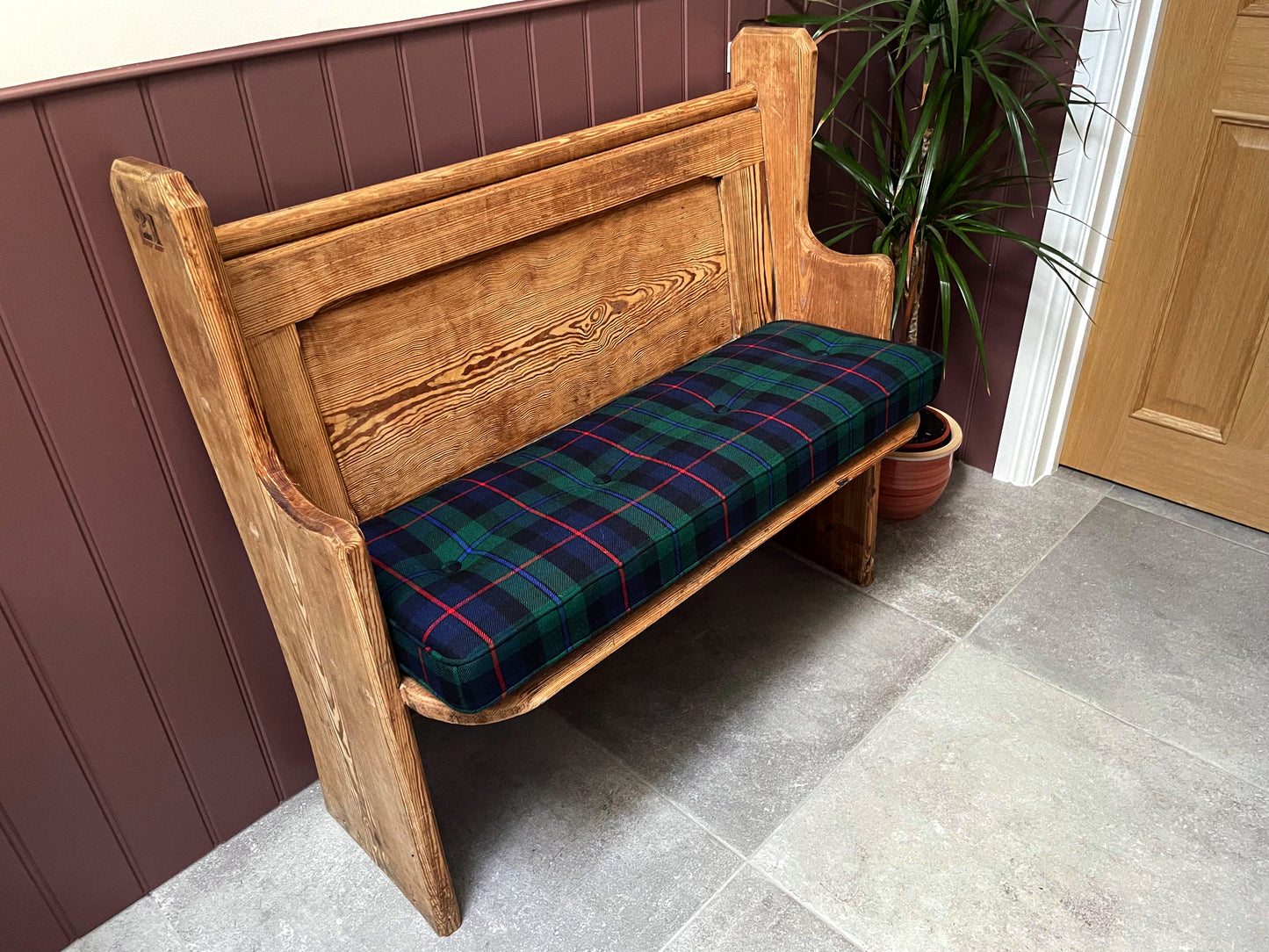 A narrow wooden church pew bench with a hand button tufted bench cushion made to measure on top. Family tartan cushions in plaid wool fabric. Buttoned and piped bench cushion for indoor bench. Navy blue, emerald green and red check patterned fabric bench cushion with firm foam and dacron wadding inside. Wipe clean natural fibre fabric bench cushion. Hard wearing and long lasting for dining bench, banquette, in built wooden seating area. Hanging bar back cushion pads with velcro and fabric ties