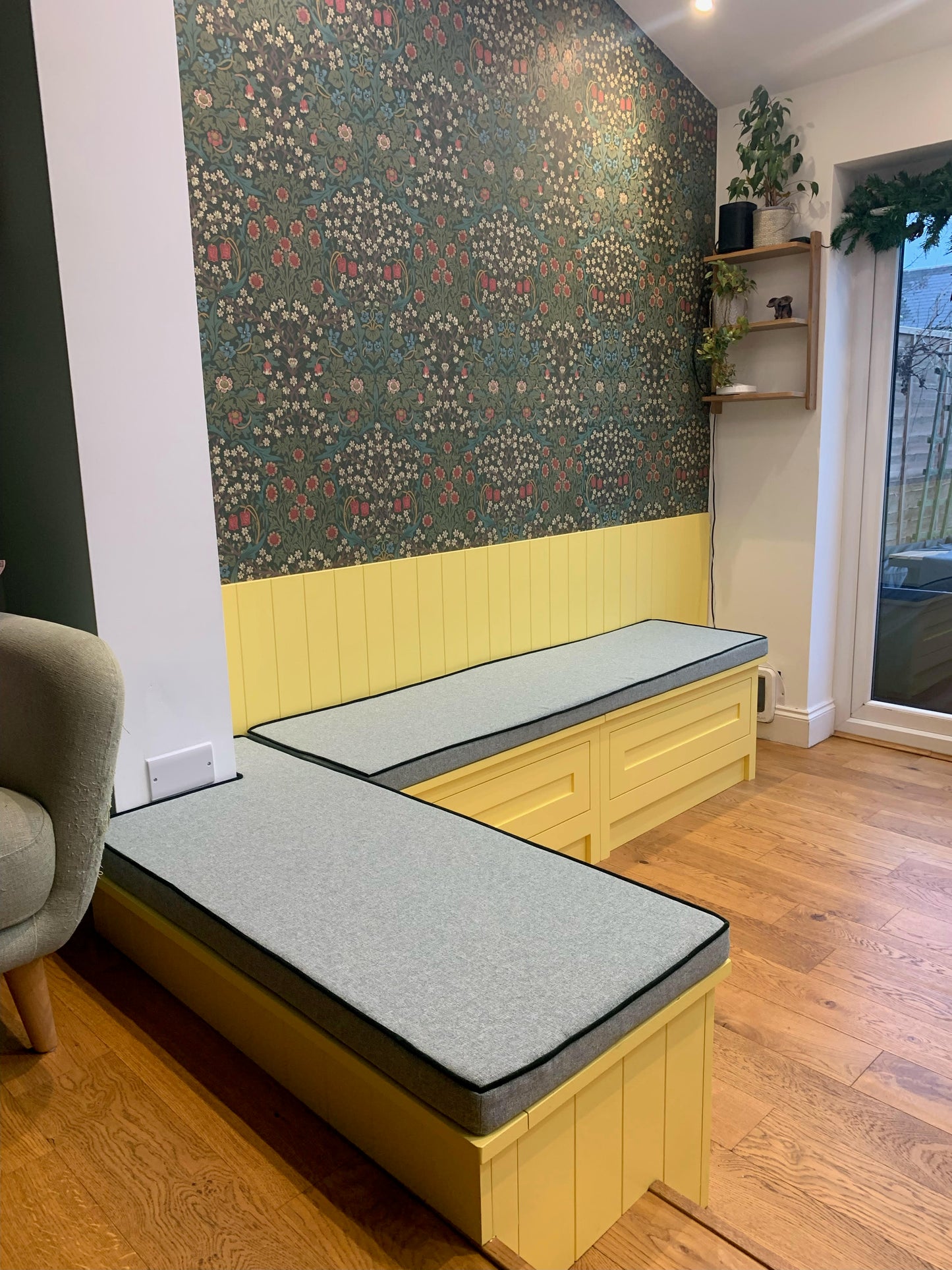 A brightly coloured dining bench area with daffodil yellow panelled walls and a dark green William Morris floral wallpaper. The wooden built in bench seating area has two custom shaped bench cushions on top. They have firm foam filling and are covered in a flecked light green upholstery fabric. The cushions have dark green contrast piping and have concealed zips. The shaped dining banquette bench cushion have cut outs to fit around a protruding wall in one corner. Paper template custom shaping