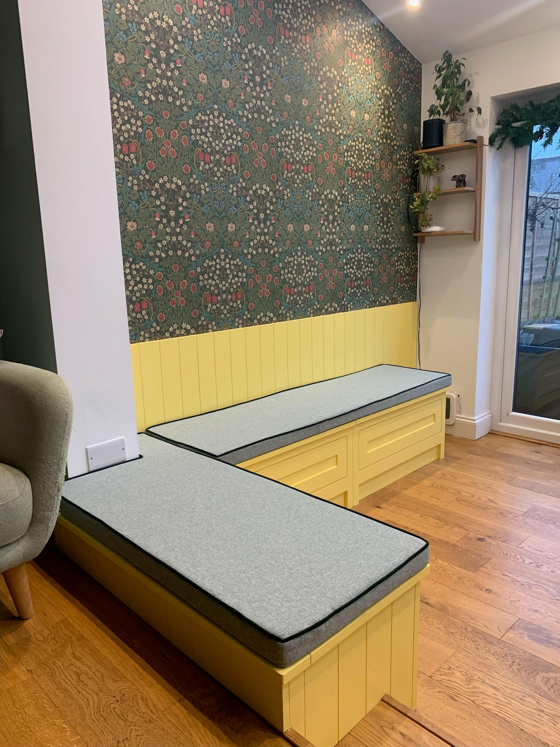 A brightly coloured dining bench area with daffodil yellow panelled walls and a dark green William Morris floral wallpaper. The wooden built in bench seating area has two custom shaped bench cushions on top. They have firm foam filling and are covered in a flecked light green upholstery fabric. The cushions have dark green contrast piping and have concealed zips. The shaped dining banquette bench cushion have cut outs to fit around a protruding wall in one corner. Paper template custom shaping