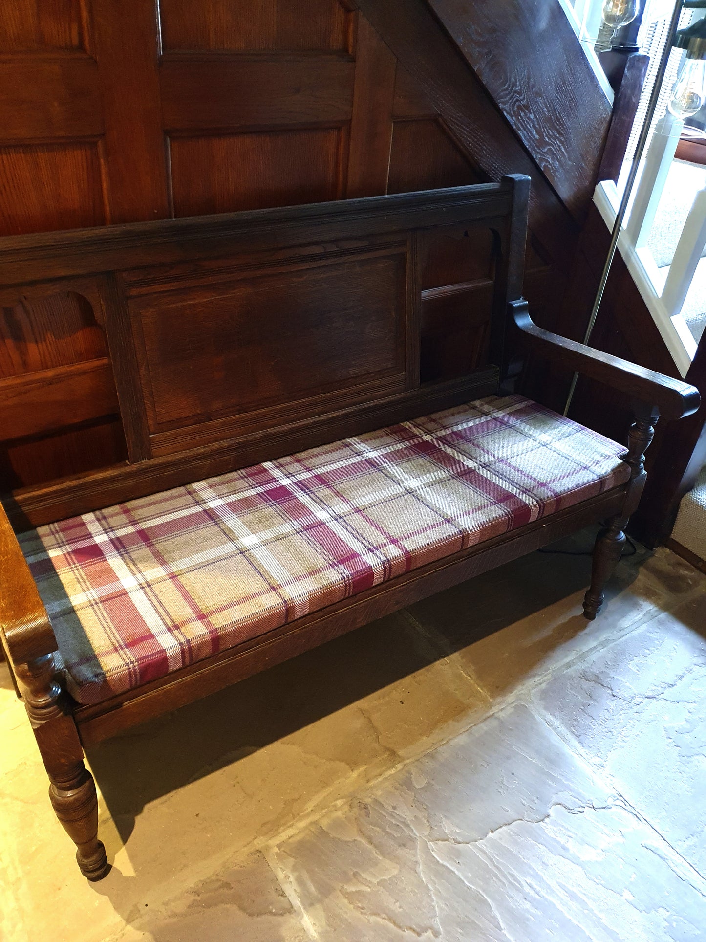 A dark wood vintage church pew seat with arms. The pew bench has a made to measure bench cushion on top in a burgundy red, green and cream check tartan pattern. The wool look fabric used for the bench cushion cover is hard wearing but soft to touch. The cushion has cut outs at the front and back of the cushion to perfectly fit around the arms and back struts of the seating bench. This wooden bench and matching bench cushion is perfect for a boot room or hallway seating.