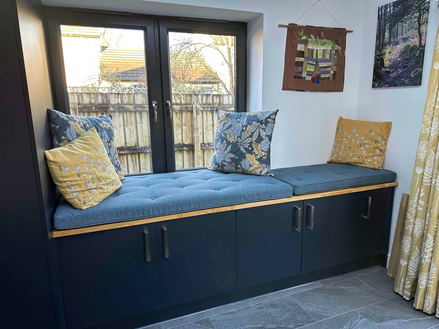 Two piped upper edge bench cushions with button tufting in a dark blue woven, hard wearing, wipe clean fabric. The storage bench seat cushions have firm foam inside with dacron and stockinette wrap. The window ledge cushions have zips at the back. Hand made blue long lasting bench cushions on a wooden, in built storage cupboard bench with dark blue painted doors. A room with a mustard yellow and teal blue colour scheme with dark wooden large windows and white painted walls. Supportive, comfy